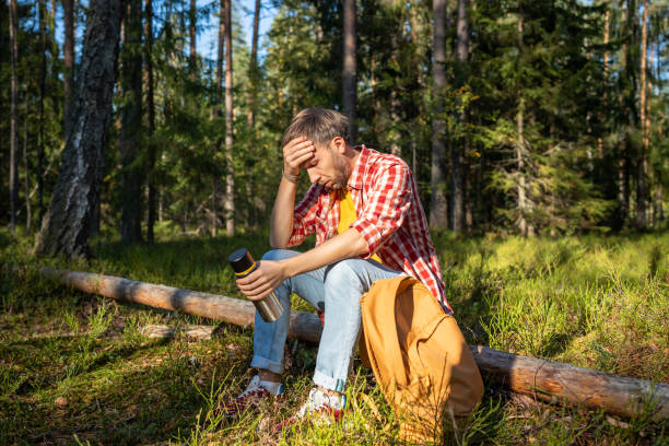 A man holds his head in pain while sitting on a log in the forest.
